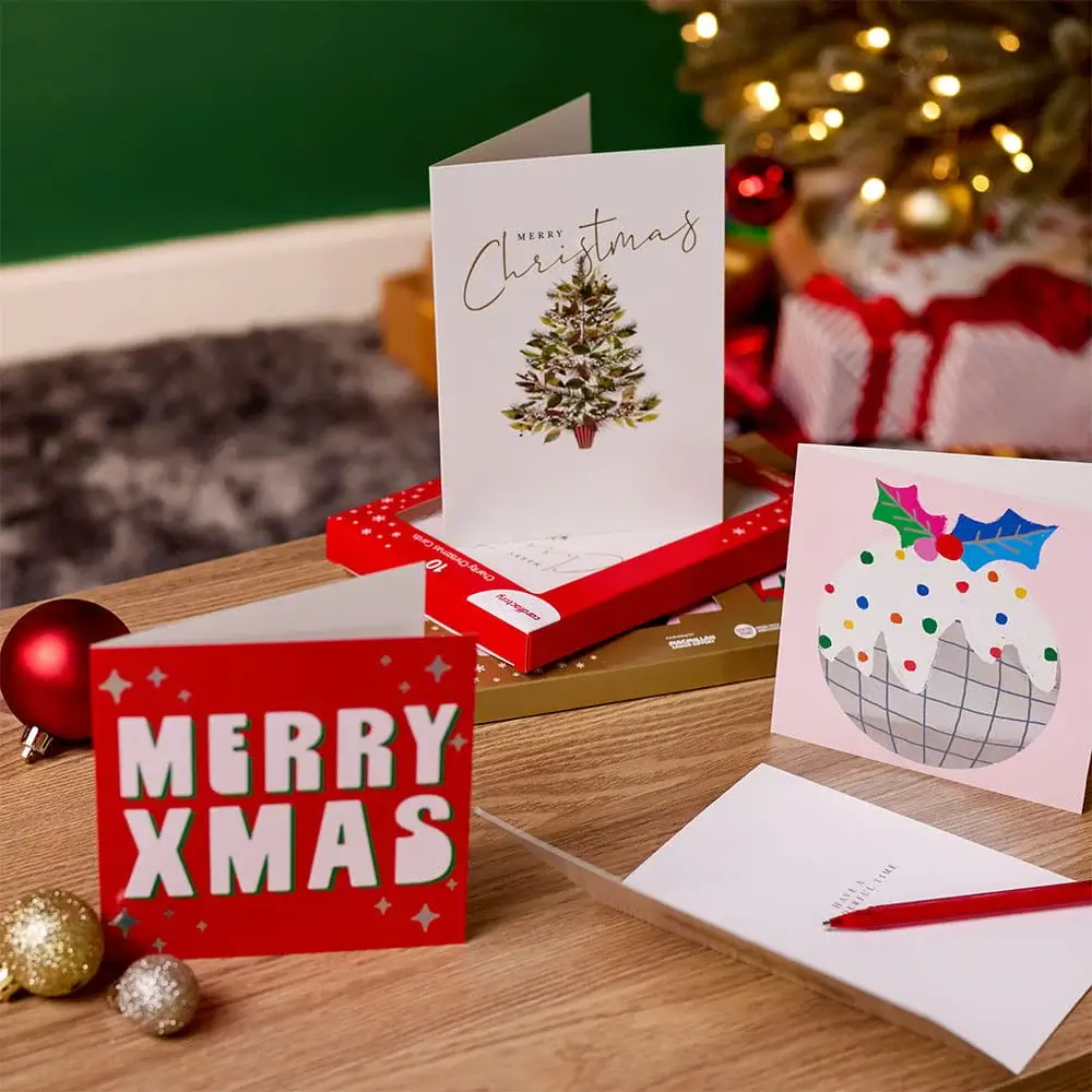 Selection of Christmas greeting cards displayed on a wooden table with festive decorations, gift boxes, baubles, and a decorated Christmas tree in the background. Selection of Christmas greeting cards displayed on a wooden table with festive decorations, gift boxes, baubles, and a decorated Christmas tree in the background.